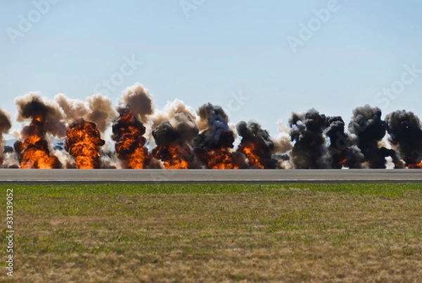 Fototapeta Multiple firey explosions with thick black smoke on an airport runway.