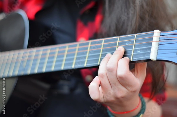 Fototapeta The neck of a classic black guitar. The girl's fingers pluck the strings. Songs with a guitar in the yard. Selective focus, close-up, background blur.