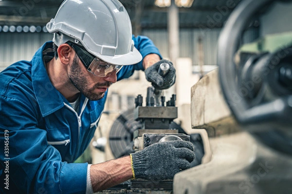 Obraz Male engineer in blue jumpsuit and white hard hat operating lathe machine.	