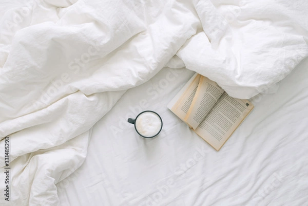 Fototapeta A cup of coffee and an open book lie on a white open bed. Top view