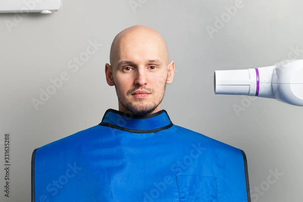 Obraz young bald man in a blue protective lead apron in an x-ray room near an x-ray apparatus for dental imaging. dentistry