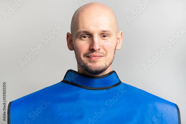 Fototapeta Smiling young bald man with a shaved head in a blue lead apron, close-up portrait on a gray background. X-ray room