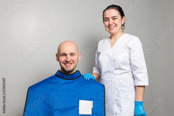 Obraz female doctor in a white medical coat and rubber gloves next to a smiling male patient sitting in a protective lead apron.