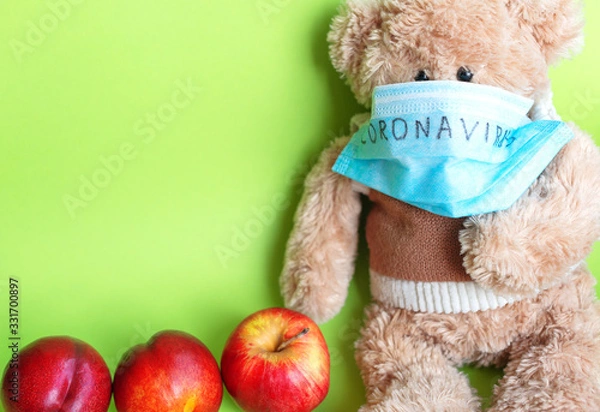 Fototapeta A soft toy bear in a protective medical mask with the inscription "coronavirus", sitting on a green background, next to apples. The concept of quarantine of schools and kindergartens, home education.