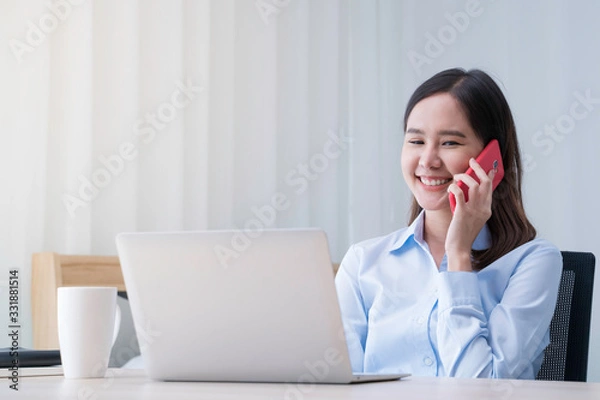 Fototapeta Attractive Asian woman works from home with laptop on desk in bedroom. Young girl smiling with happiness face while talking on phone. Telecomuting, freelancer and modern lifestyle concept.