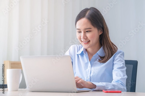 Fototapeta Attractive Asian woman works from home with laptop on desk in bedroom. Young girl smiling with happiness face while reading at computer screen. Telecomuting, freelancer and modern lifestyle concept.