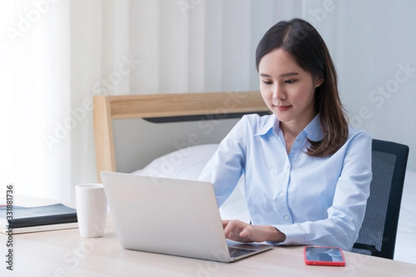 Fototapeta Attractive Asian woman works from home with laptop on desk in bedroom. Young girl smiling with happiness face while reading at computer screen. Telecomuting, freelancer and modern lifestyle concept.
