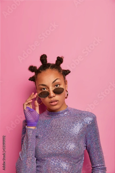 Fototapeta Self confident attractive female has funny hairstyle with buns, keeps hands on shades, wears glittering jumper, looks directly at camera, poses against pink pastel wall, empty space. People, style