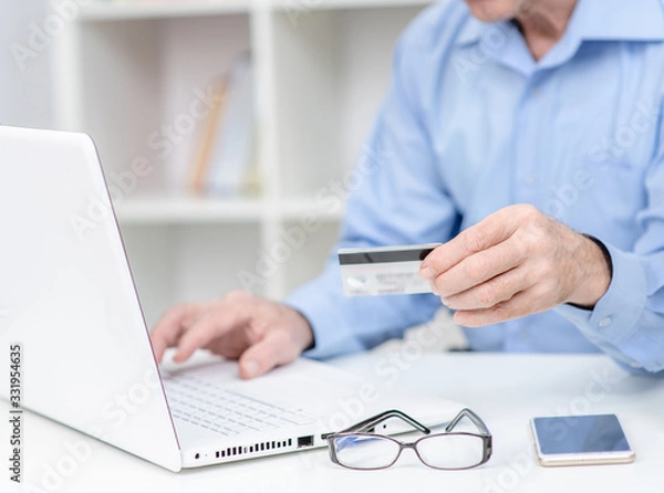 Fototapeta Senior man holds credit card and uses laptop. Online shopping
