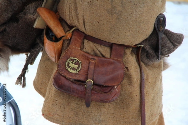 Fototapeta Medieval leather historical waist bag with a copper plaque in the shape of a reindeer hangs on a mans belt in medieval clothes with an ax behind his belt
