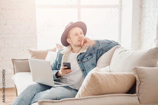 Fototapeta Young man dreams startup business, works home in living room with laptop. Chatting with friends via online chat rooms phone, light background, sunlight from window