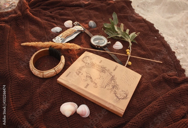 Fototapeta Notepad, compass, knife, olive branch, spikelet, bone bracelet and shells on a brown bedspread in the sand.