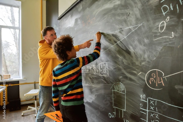 Obraz The great aim of education is not knowledge but action. Young male science teacher explaining main rules and formulas in physics and mechanics. A boy is drawing on the blackboard