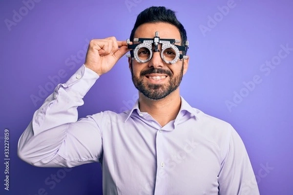 Fototapeta Young handsome optical man with beard wearing optometry glasses over purple background with a happy face standing and smiling with a confident smile showing teeth