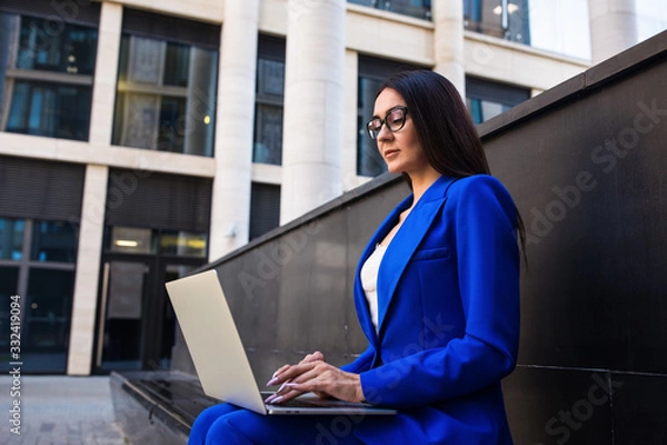Obraz Serious woman executive director online banking via laptop computer while sitting outdoors near company building. Female government worker using notebook gadget for chat in internet