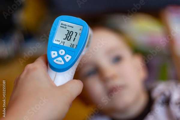 Fototapeta A hand with an electronic thermometer is aimed at the child, on the thermometer the temperature is 38.7, measurement of body temperature by an electronic thermometer