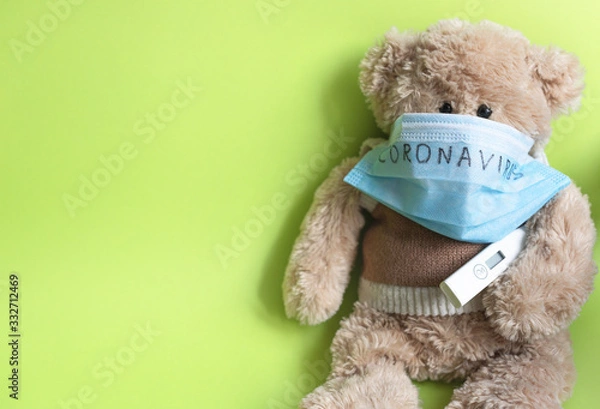 Fototapeta Soft toy bear in a protective medical mask with the inscription "coronavirus" and a thermometer, sitting on a green background. The concept of home education from the COVID-19 quarantine