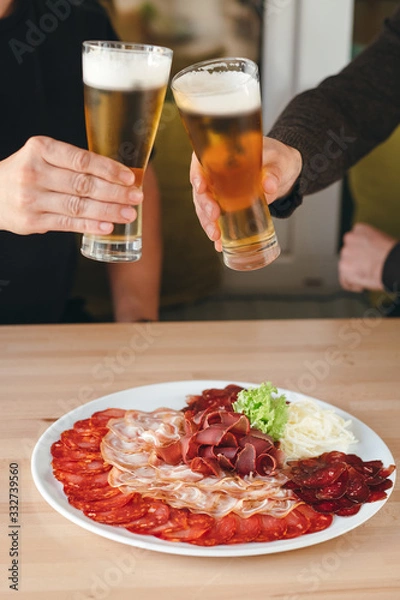 Fototapeta Cold smoked meat plate with salami, pastrami and cheese on a plate and two hands holding beers.