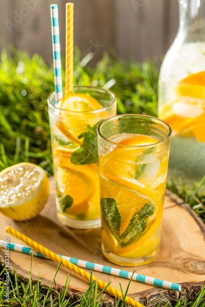 Obraz cold lemonade drink with orange and mint in tall glass on green grass lawn wooden background