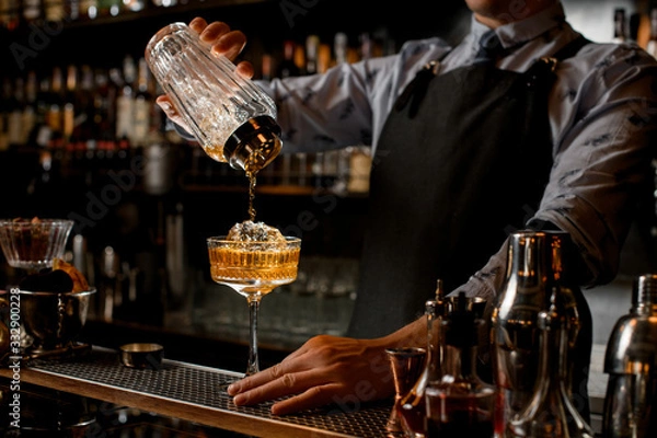 Fototapeta Professional bartender in black apron pours drink from shaker into glass.
