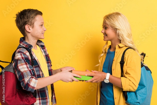Fototapeta side view of smiling schoolkids holding books on yellow background