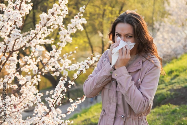 Fototapeta Young pretty woman sneezing in a park with blooming trees in spring. Pollen Allergy Symptoms