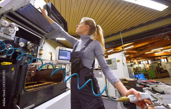 Fototapeta A young woman works as a computerized  numerical control technician. She is seen at her workplace wearing a blue overall. 