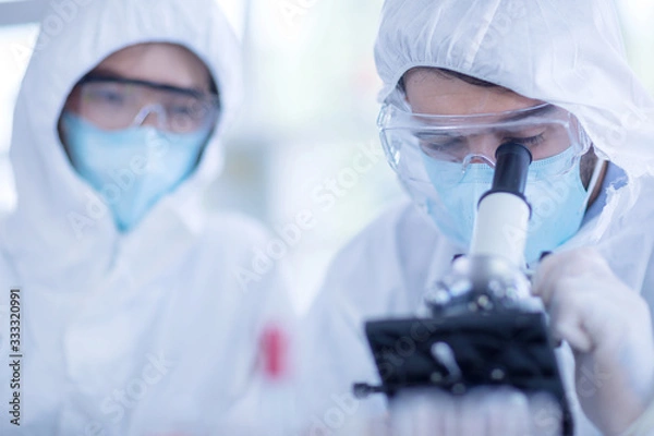 Obraz A male scientist wearing a mask and looking through the microscope in the laboratory, Concept, The scientists wear white protective clothing is experimenting with drug anti- Coronavirus, Covid19