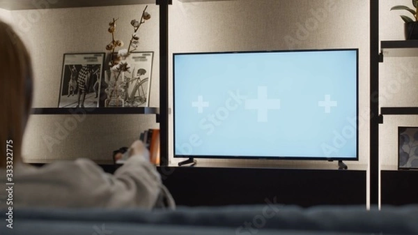 Fototapeta Over shoulder close up view of young woman changing channels with a TV remote control on a green white chroma key screen mock-up with track points sitting on sofa relaxing in cozy home warm lights