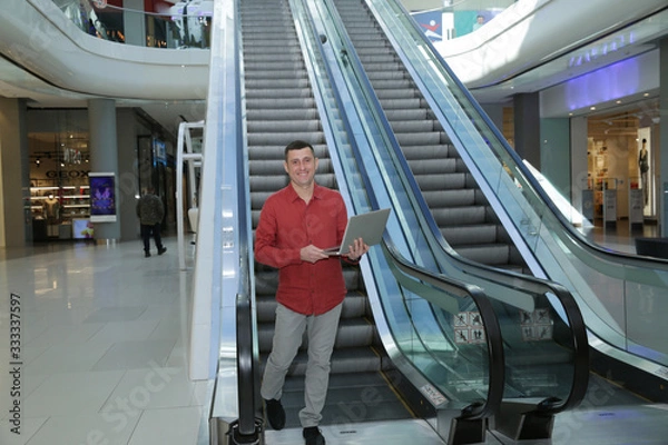 Fototapeta Young handsome businessman in red shirt on an escalator with an open laptop in his hands in a business center of a shopping center or airport.