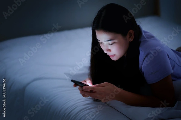 Fototapeta Asian young woman lying on the white bed and playing smartphones during night time. She is chatting with her friend. Using phone in low light is impact to eyes. Health and social Concept