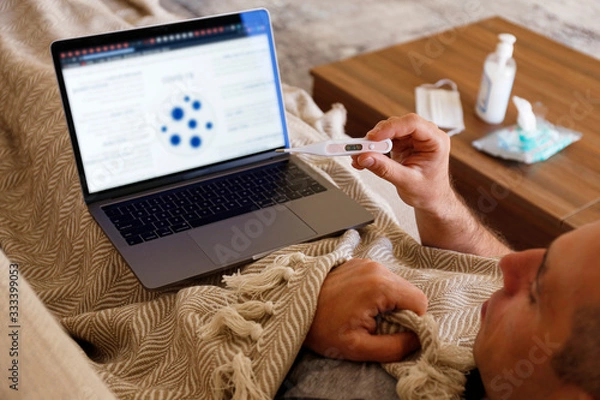 Fototapeta Close up shot of adult man working at home due to coronavirus quarantine concept. Male sitting on couch with laptop, measuring body temperature. Antiseptic gel and wet wipes on background, copy space.