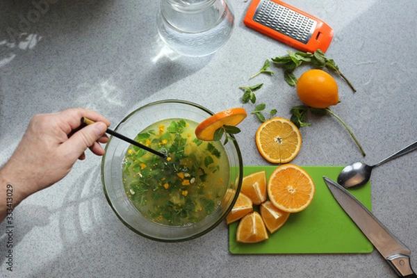 Fototapeta The process of making homemade lemonade. Lemon, orange, mint are on the gray table. A man's hand mixes ingredients in a transparent bowl. Concept of healthy food, lifestyle and diet. Selective focus.