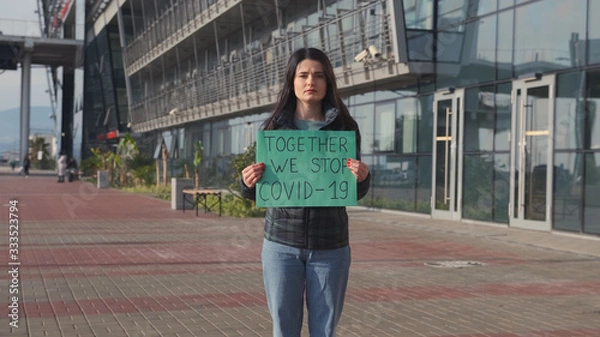 Obraz Pandemic, portrait of a young woman wearing a protective mask holding a poster with the inscription "TOGETHER WE STOP CAVID-19". The concept of health and protection from the pandemic COVID-19.