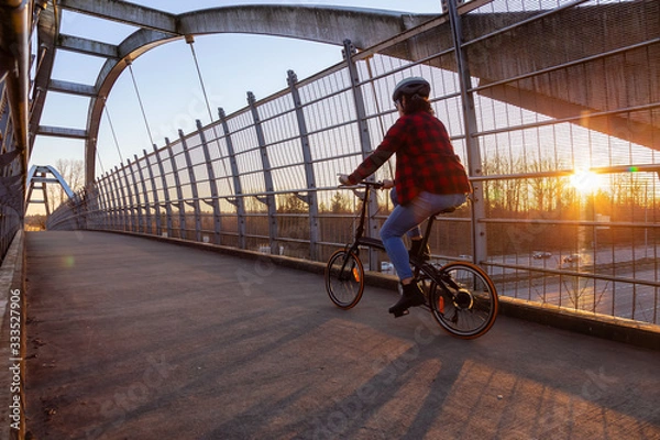 Fototapeta Caucasian Woman Riding a Bicycle on a Pedestrian Bridge over the Highway during a sunny sunset. Taken in Surrey, Vancouver, British Columbia, Canada.