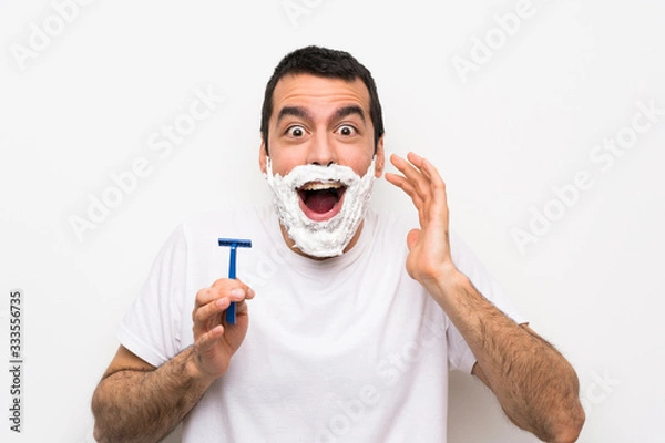 Fototapeta Man shaving his beard over isolated white background with surprise and shocked facial expression
