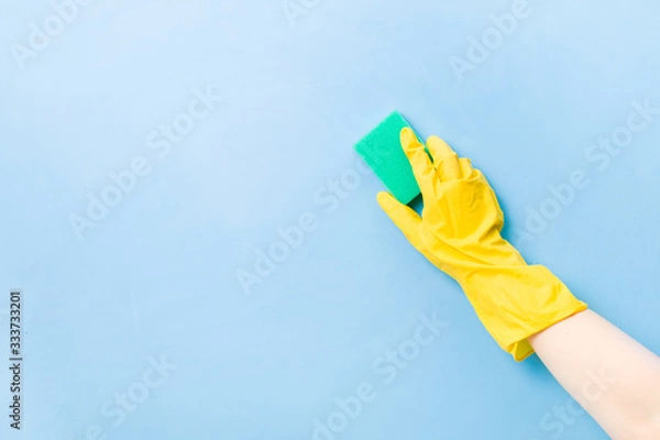 Obraz hand in a yellow rubber glove holds a green sponge for washing dishes and cleaning, blue background copy space, cleaning concept, household chores, spring cleaning