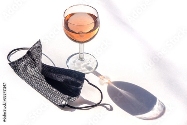 Fototapeta Medical face mask black next to a glass of rose wine on a white background copy-space. Concept of social activities during pandemic and crown virus quarantine.