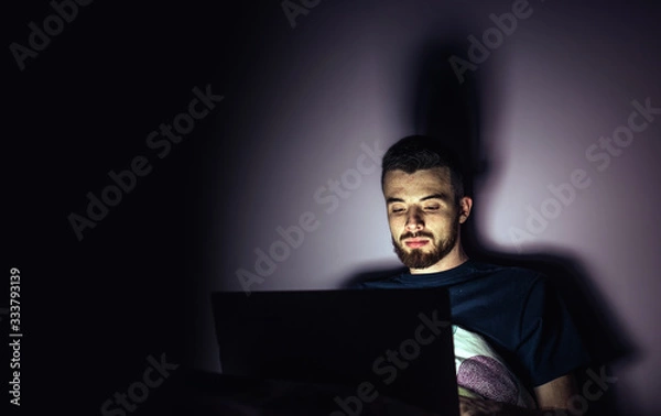 Fototapeta Young man lying on bed and look at laptop screen. Calm concentrated guy working overtime playing games or entertain himself. Night room. Isolated over dark background.