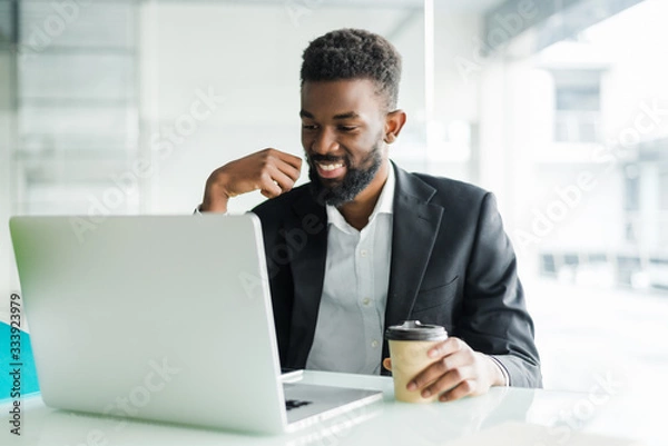 Fototapeta Thoughtful African American businessman using laptop, pondering project, business strategy, puzzled employee executive looking at laptop screen, reading email, making decision at office