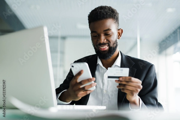 Fototapeta African American man paying with credit card online while making orders via mobile Internet making transaction using mobile bank application.