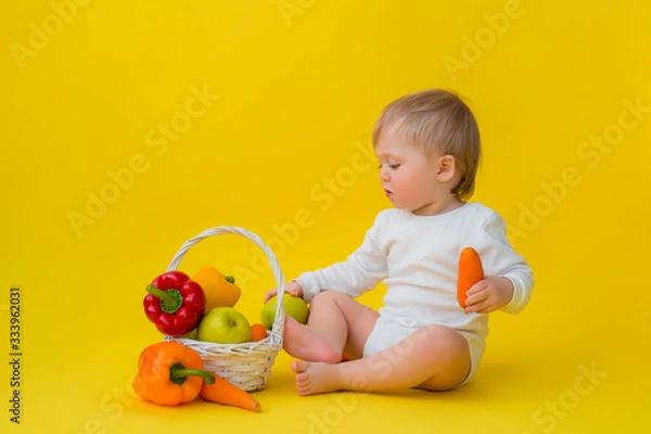 Fototapeta baby in a white bodysuit with vegetables, sitting on a yellow background. healthy food for the child, space for text