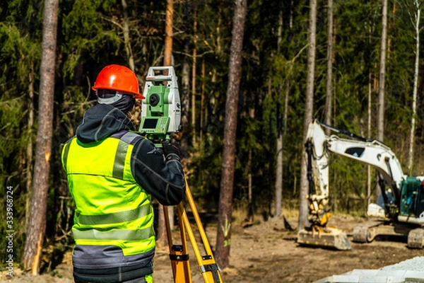 Fototapeta Surveyor engineer with equipment (theodolite or total positioning station) on the construction site of the road or building with construction machinery background