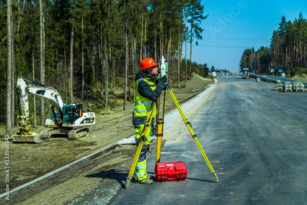Fototapeta Surveyor engineer with equipment (theodolite or total positioning station) on the construction site of the road or building with construction machinery background