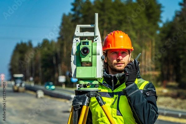 Fototapeta Surveyor engineer with equipment (theodolite or total positioning station) on the construction site of the road or building with construction machinery background
