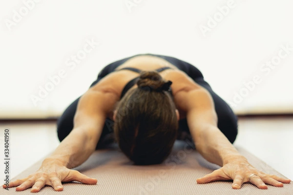 Fototapeta Yogi woman practices child pose in bright studio. Focus on hands of lady in balasana on purple mat. Healthy lifestyle, resting exercise, workout, sportswear, yin yoga concepts. Vintage effect applied