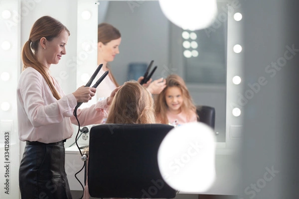 Fototapeta The work of the stylist on hairstyles in a bright salon, the girl makes curls and braids braids of a small beautiful model,