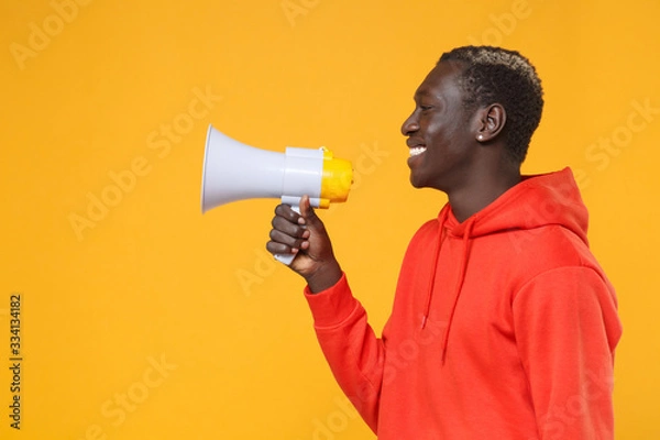 Fototapeta Side view of smiling young african american man guy in red streetwear hoodie posing isolated on yellow background studio portrait. People emotion lifestyle concept. Mock up copy space. Hold megaphone.