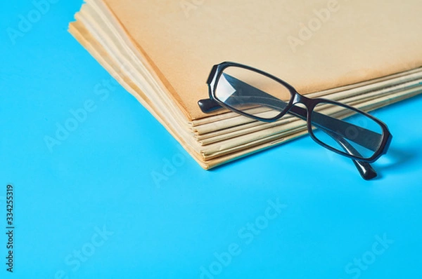 Fototapeta Heap of blank magazines, newspapers or some documents and modern glasses on blue desk. Business concept. Workplace of journalist or secretary
