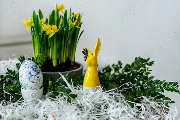 Fototapeta Egg and daffodils. Easter egg decoration, next to yellow porcelain rabbit and yellow narcissi growing in pot, grey background. Easter holiday composition.